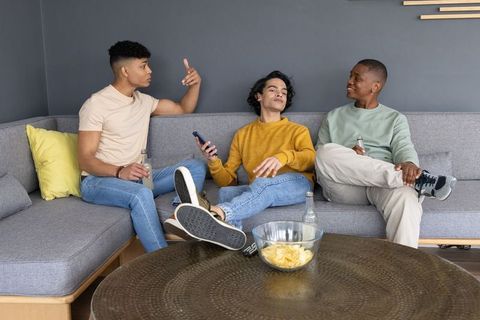 Young diverse friends relaxing on l-shaped gray sofa, chatting and checking smartphone