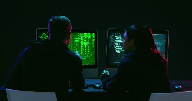 Caucasian couple working on computers in a dark room. They are focused on coding or cybersecurity tasks, illuminated by screen light.