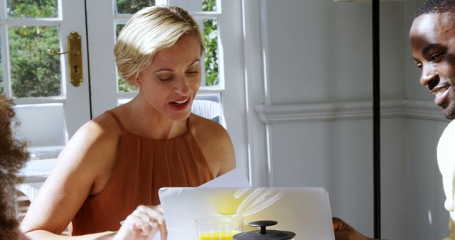 Woman working on laptop at home while interacting with friend. Sunlight streaming through window. Suitable for concepts such as remote work, home office, friendship, collaboration, casual work environment, work-life balance.