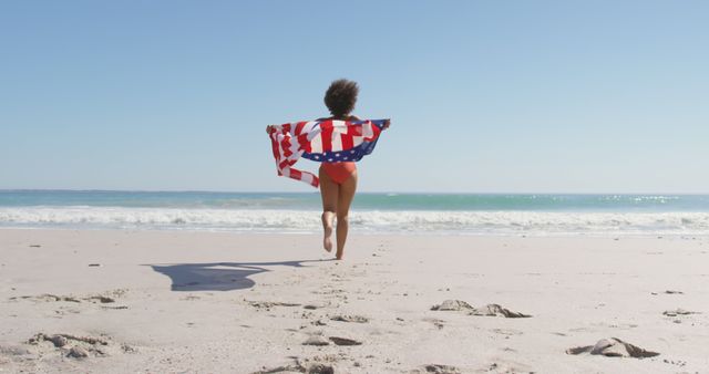 Woman running on sandy beach holding American flag with ocean in background. Perfect for promoting summer vacations, USA holidays, patriotic events, beach tourism, travel brochures, and Fourth of July celebrations.