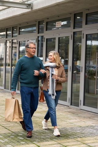 Walking arm-in-arm senior couple enjoying urban shopping stroll with coffee and paper bags