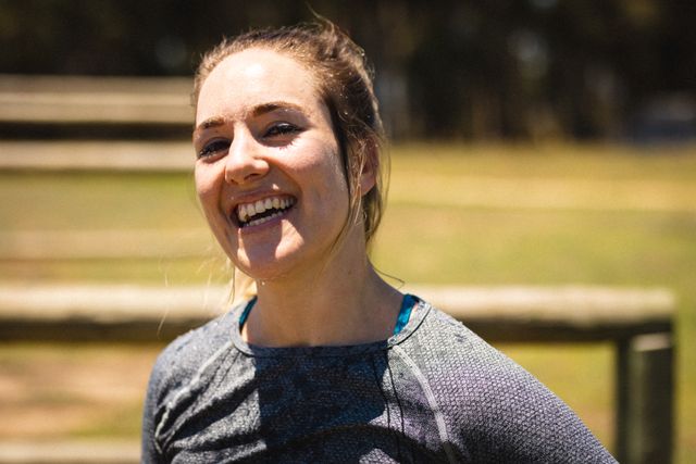 Close-up of a smiling Caucasian mid adult woman participating in an outdoor bootcamp training session. She is wearing sports clothing and appears to be enjoying the sunny weather. This image is perfect for use in fitness and health-related promotions, advertisements for outdoor training programs, or articles about active lifestyles and exercise.