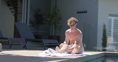 Shirtless man applying lotion to feet while sitting on pool deck towel wearing sunglasses