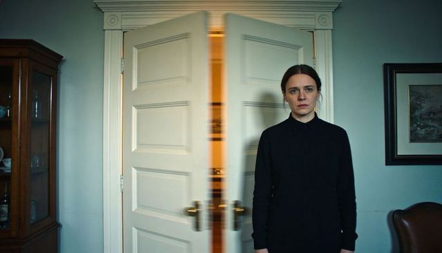 Woman standing by swinging white doors revealing warm interior light in moody blue room