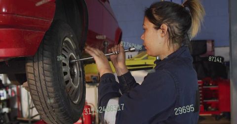 Female mechanic tightening wheel with breaker bar under lifted red sedan in auto repair garage