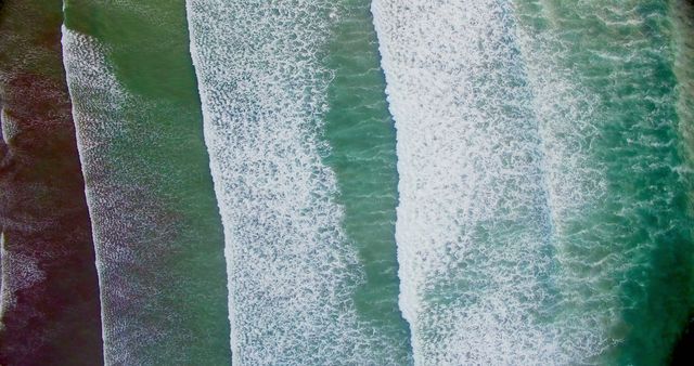 Image shows top-down view of ocean waves crashing onto sandy shore, creating white foam patterns. Ideal for use in travel brochures, websites promoting beach vacations, coastal environmental campaigns, landscape calendars, and decorative prints.