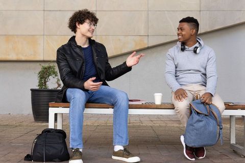 College friends conversing on campus bench, backpacks and coffee, casual urban lifestyle