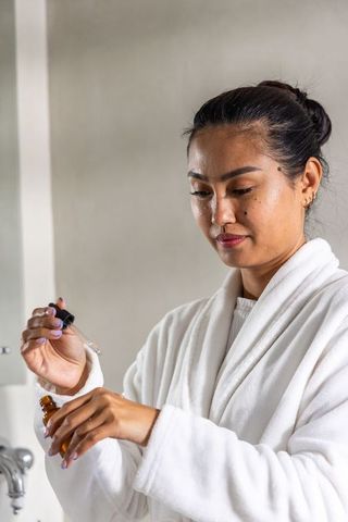 Asian woman applying serum with pipette at sink wearing white robe, skincare routine