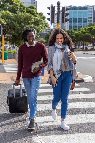 Friends walking across city crosswalk pulling suitcase and checking devices