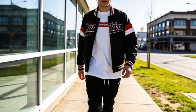Young man walking urban sidewalk past glass facade wearing black varsity jacket and red box logo tee