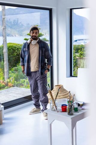 Artist standing by window in waterfront home studio wearing beanie and headphones