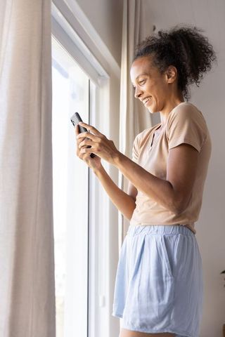 Smiling woman texting beside sunlit window in minimalist home, relaxing lifestyle moment