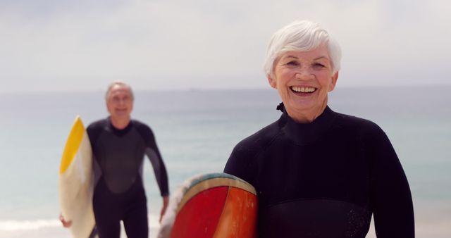 A senior Caucasian woman smiles brightly in the foreground holding a surfboard, with a senior man in a wetsuit approaching from behind, both at the beach, with copy space. Their active lifestyle and joy in surfing reflect a positive approach to aging and fitness.