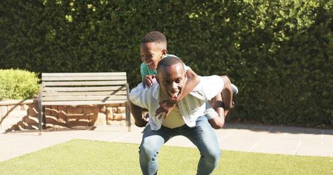 Father giving piggyback ride to laughing son on sunny backyard lawn, playful family bonding