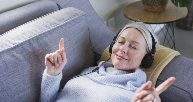 Relaxed senior caucasian woman in living room lying on sofa, wearing headphones. retirement lifestyle, at home with technology.