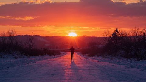 Silhouetted person embracing winter sunset on snowy rural road with long shadow