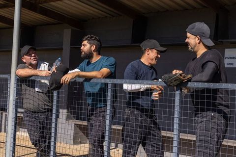 Baseball teammates leaning on dugout railing chatting and adjusting glove on sunny day