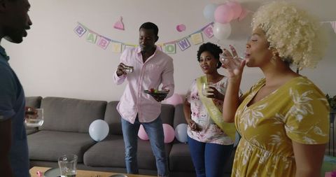 African American friends celebrating home baby shower with pastel balloons, banner, drinks