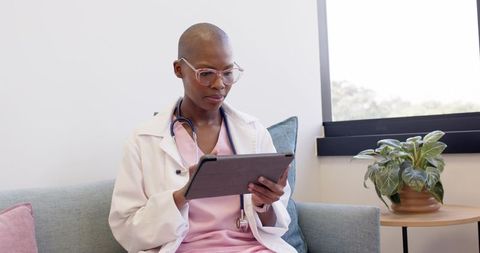 Black female doctor reviewing tablet on clinic sofa wearing pink scrubs and stethoscope