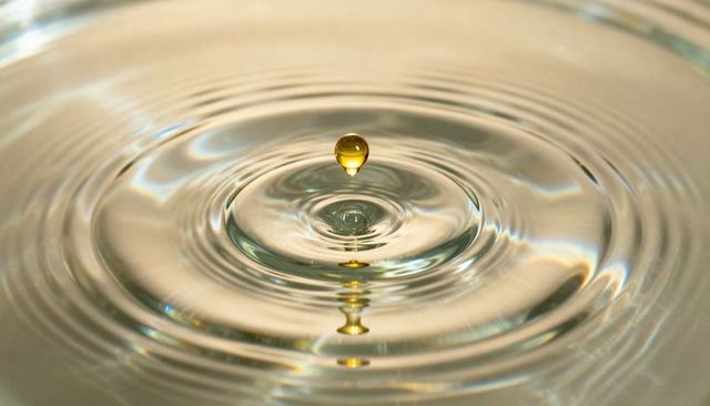 Golden water droplet hovering above concentric ripples with reflected column, macro