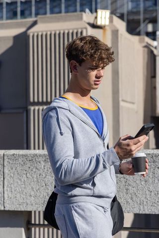 Young man standing on rooftop using smartphone holding coffee in gray hoodie and sweats