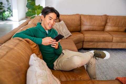 Asian man relaxing on leather sectional texting on smartphone in sunlit living room