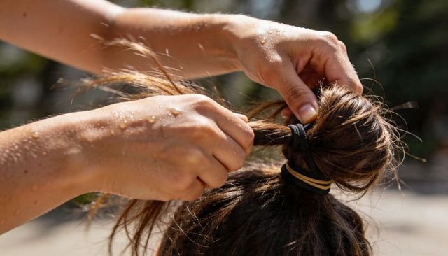 Hands gathering sunlit dewy hair into messy topknot with black and yellow elastics