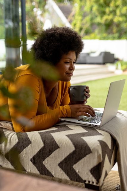 Smiling african american mid adult woman with coffee cup using laptop while lying on couch. unaltered, wireless technology, drink, lifestyle and domestic life concept.