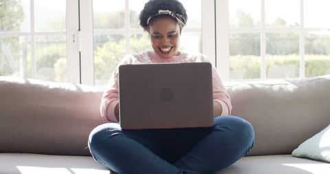 Content Woman Smiling While Using Laptop on Couch in Sunlit Room