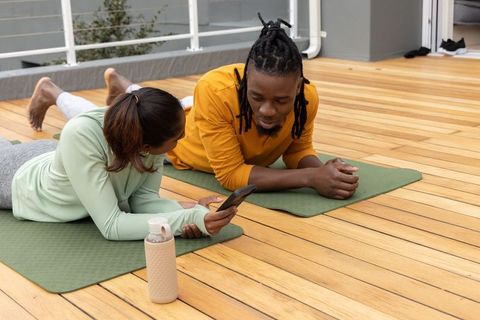 Couple practicing yoga on rooftop deck checking workout app relaxing on green mats
