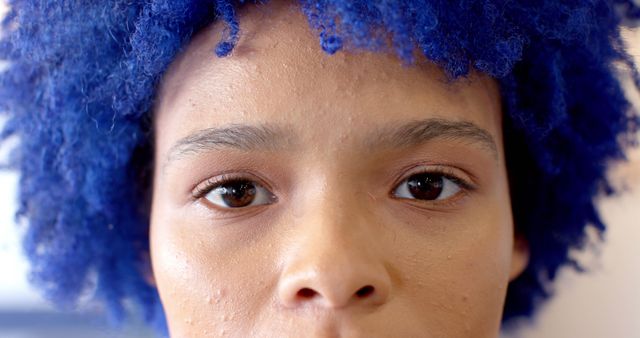 This image shows a close-up portrait of a young person with vibrant blue curly hair and a serious expression. The photo emphasizes their unique hairstyle and skin texture, making it suitable for topics on self-expression, individuality, fashion, and edgy styles. Ideal for use in magazines, social media, and mental health awareness campaigns focusing on personal identity and confidence.