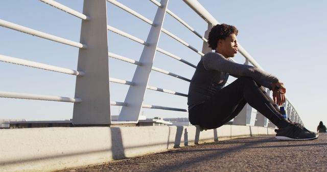 Young athlete sitting on the ground of an urban bridge, taking a break from exercise. Background features blue sky and geometric lines of the bridge. Suitable for fitness, sportswear promotional materials, lifestyle blogs, and advertisements focusing on active and healthy living in urban environments.
