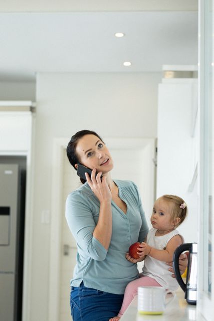 This image depicts a busy mother multitasking at home, talking on a smartphone while holding her baby daughter on the kitchen counter. It is ideal for use in articles or advertisements related to parenting, work-life balance, remote work, and family life during quarantine or lockdown. It can also be used in blogs or social media posts about modern motherhood and the challenges of balancing work and family responsibilities.