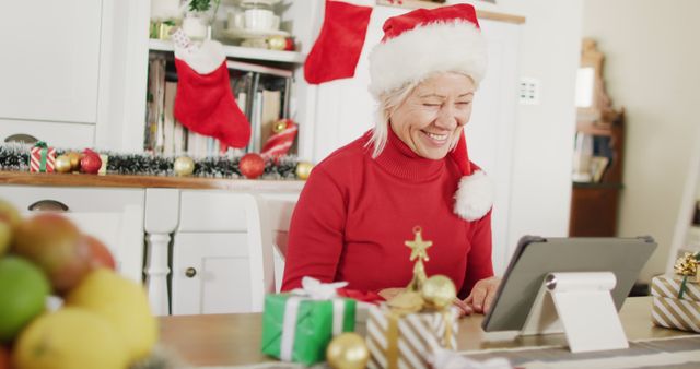 Happy senior caucasian woman wearing santa claus hat, using tablet for image call. Spending quality time at home alone at christmas.