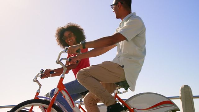 Young biracial couple riding bicycles along a sunny beach promenade, enjoying quality time together. Their smiles convey happiness and connection, perfect for illustrating themes of recreation, friendship, and active lifestyle. Ideal for advertisements promoting a feel-good summer atmosphere, romantic getaways, or leisure wear campaigns.