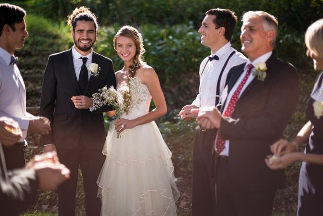 Bride and groom standing with guests in a park, celebrating their wedding day. Ideal for use in wedding invitations, bridal magazines, event planning websites, and social media posts about weddings and celebrations.