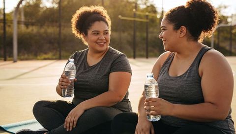 Two women sharing post-workout break on sunlit outdoor court holding water bottles