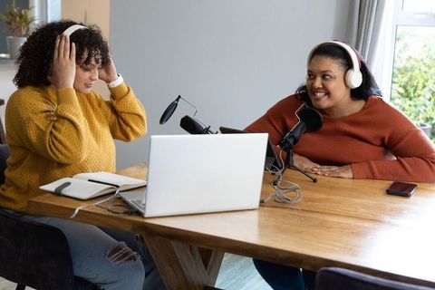 African American and Indian women podcasting from home recording with mics and laptop