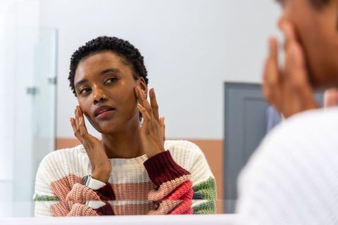African american woman checking skin in bathroom mirror touching face wearing smartwatch
