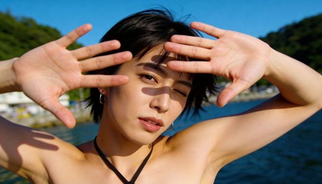 Sunlit asian woman shielding eyes with hands at harbor, halter strap and hoop earrings