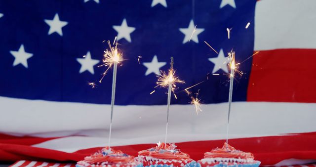 Sparklers light up atop red, white, and blue frosted cupcakes in front of an American flag, suggesting a celebration of a patriotic holiday. The image captures a festive Fourth of July or Memorial Day theme, with copy space.