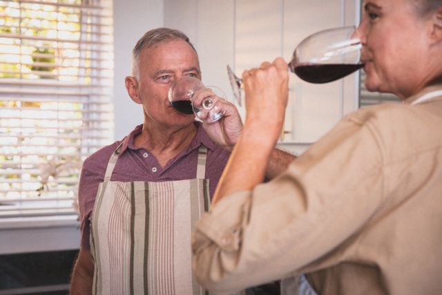 Senior couple enjoying red wine in their kitchen, wearing aprons and spending quality time together. Ideal for use in articles or advertisements about retirement, home life, quarantine activities, and senior lifestyle.