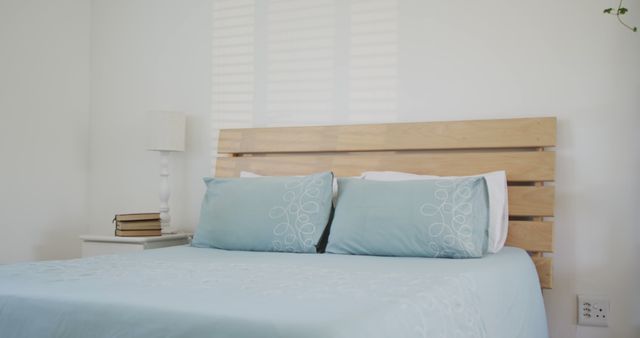 This modern minimalist bedroom features a wooden headboard, light blue pillows, and bedding that fosters a calming and serene atmosphere. A bedside table with books and a white lamp add a touch of practicality. The clean white walls and uncluttered space make it ideal for use in articles or posts about home decor, interior design, or minimalist living.