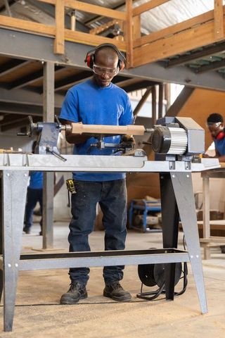 Woodworker turning wooden spindle on lathe in carpentry workshop wearing safety gear