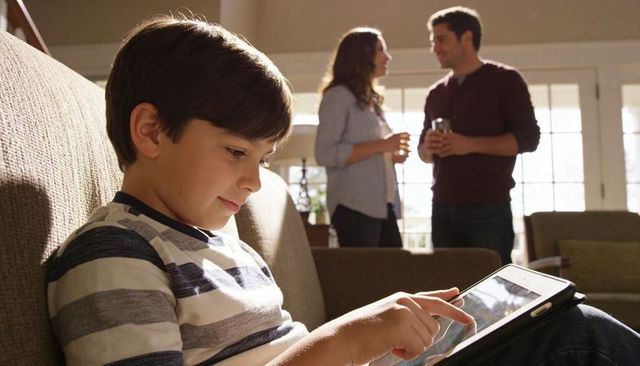 Young boy using tablet on sofa while parents chatting in sunlit cozy living room relaxed family mome