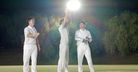 Night cricket teammates celebrating under bright floodlight on grassy pitch, players in white kits