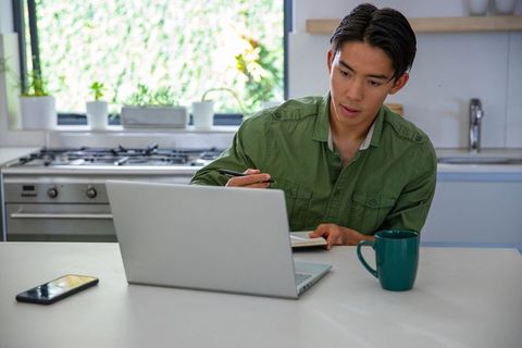 Asian man working from home at kitchen counter with laptop and notebook, productivity