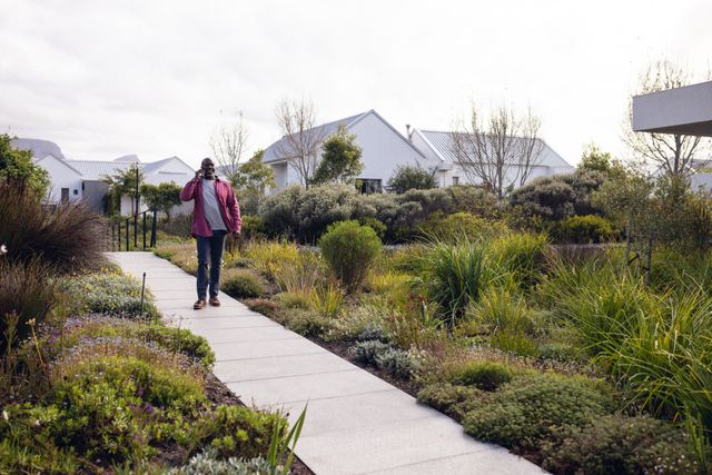 African American man walking in a lush garden while talking on the phone. Appears happy and engaged in conversation. Can be used for lifestyle, communication, and outdoor activity themes. Suitable for promoting modern living, residential areas, and nature-related content.