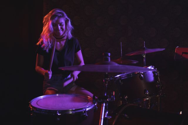 Young female drummer passionately playing drums on stage in a dimly lit nightclub. Ideal for use in articles or advertisements about live music, nightlife, female musicians, and entertainment events.