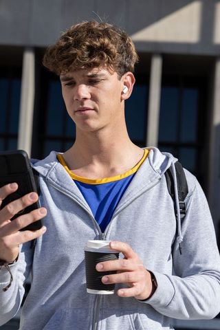 College student checking smartphone while holding coffee and wearing earbuds on campus